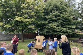 children around the Future Machine, some turning the handle, three children dressed up with a hat, a scroll and a bell, other children watching, Rachel talking and Caroline taking a photograph