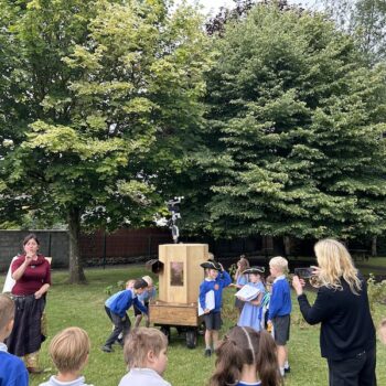 children around the Future Machine, some turning the handle, three children dressed up with a hat, a scroll and a bell, other children watching, Rachel talking and Caroline taking a photograph