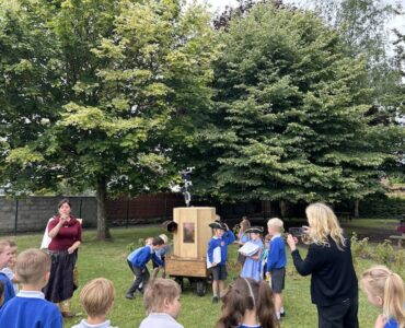 children around the Future Machine, some turning the handle, three children dressed up with a hat, a scroll and a bell, other children watching, Rachel talking and Caroline taking a photograph