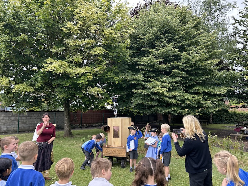 children around the Future Machine, some turning the handle, three children dressed up with a hat, a scroll and a bell, other children watching, Rachel talking and Caroline taking a photograph