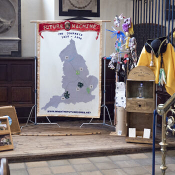 An installation with a banner with a map of England and Future Machine in red silk on the top, a sign, a wooden panel with video playing through a porthole, ittle creatures on a pole made from rubbish a wooden cabinet with a sign saying 'Cabinet of Curious Places' attached to the top, a yellow costume, a black hat and a brass bell