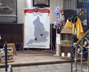 An installation with a banner with a map of England and Future Machine in red silk on the top, a sign, a wooden panel with video playing through a porthole, ittle creatures on a pole made from rubbish a wooden cabinet with a sign saying 'Cabinet of Curious Places' attached to the top, a yellow costume, a black hat and a brass bell
