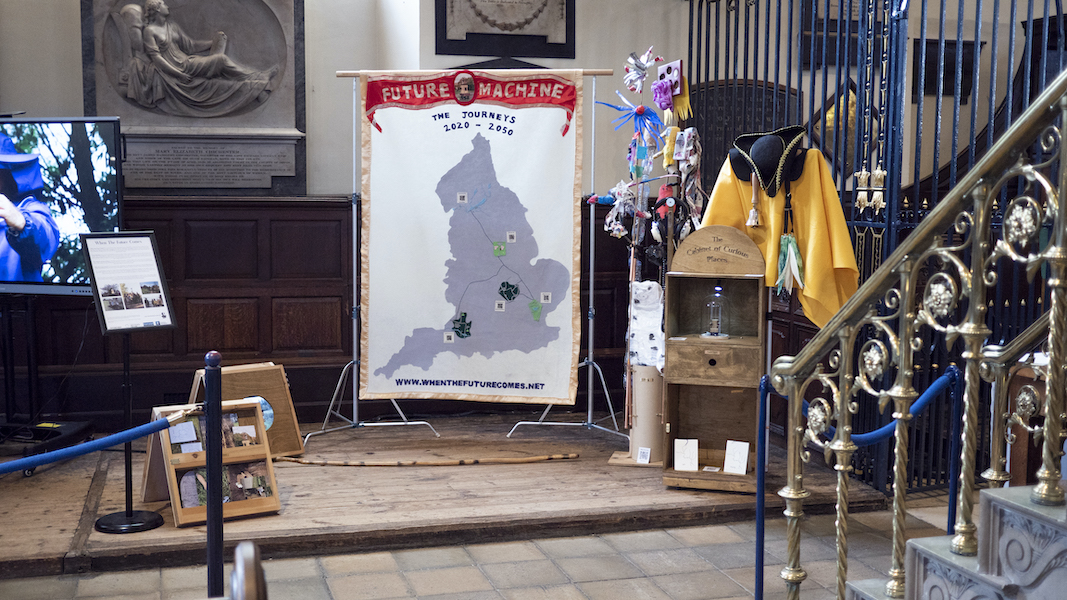 An installation with a banner with a map of England and Future Machine in red silk on the top, a sign, a wooden panel with video playing through a porthole, ittle creatures on a pole made from rubbish a wooden cabinet with a sign saying 'Cabinet of Curious Places' attached to the top, a yellow costume, a black hat and a brass bell