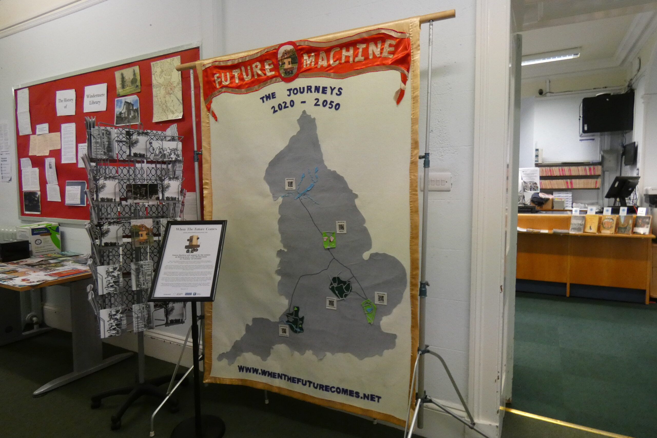 A large banner hanging against a wall, next to a doorway to the library. On the banner is a grey map of England, the words 'The Journeys 2020-2050' and symbols and QR codes on the map. A small sign in front with an image of Future Machine on the top.