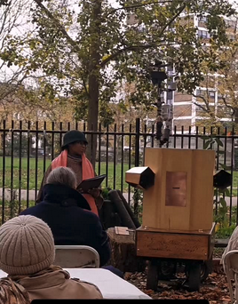 People sitting watching Esi talking next to Future Machine, with a tree behind.