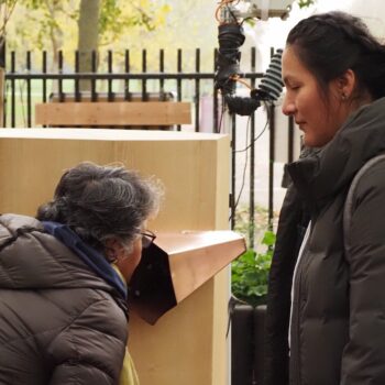 a woman talking into a copper trumpet on Future Machine's side with another woman watching