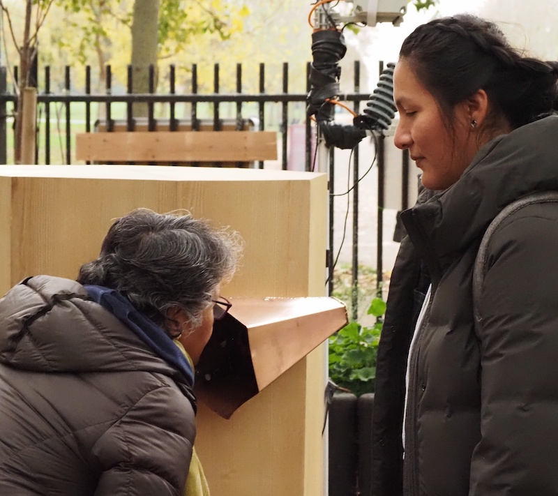 a woman talking into a copper trumpet on Future Machine's side with another woman watching