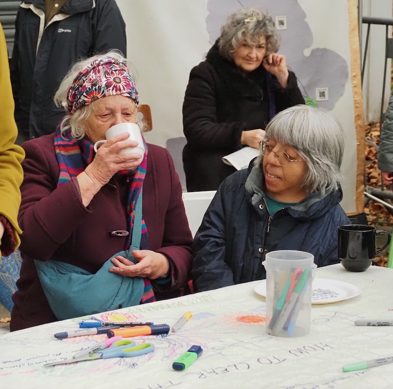 Two women sitting down at a table, one is drinking tea. There are pens and drawings on the table.